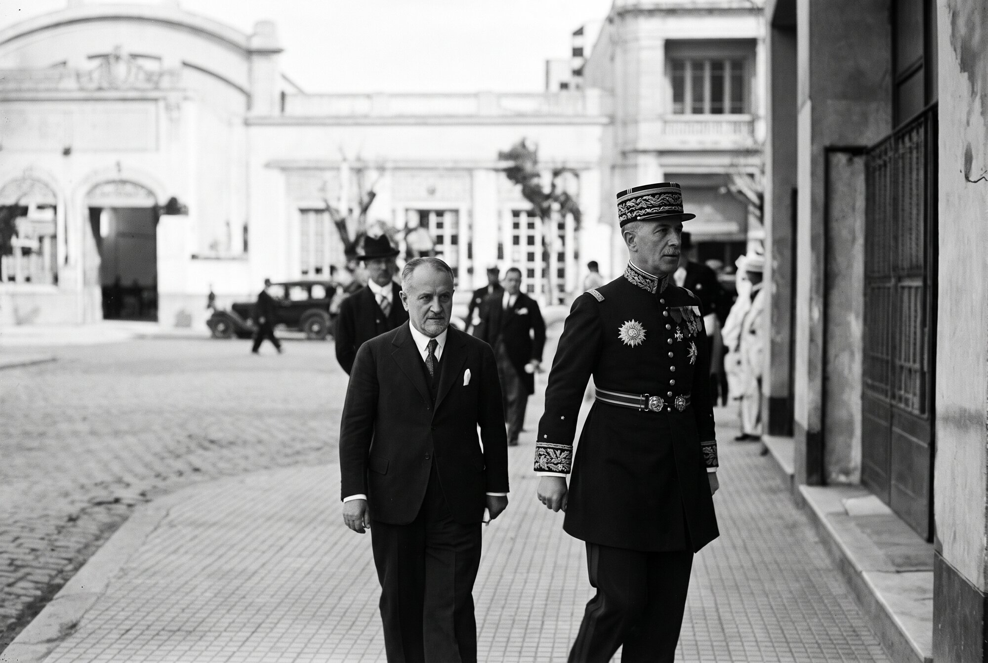 Le général Noguès en grand uniforme et François Cogné marchant sur le trottoir de l’avenue Poeymirau le long de l’Assayag ; en face, le bâtiment de la Société Hamelle