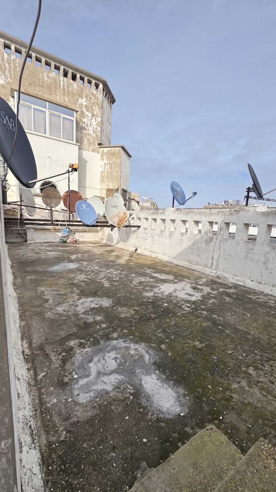 The eighth-floor drying terrace in 2026: bare concrete floor, satellite dishes lined up along the pierced parapet
