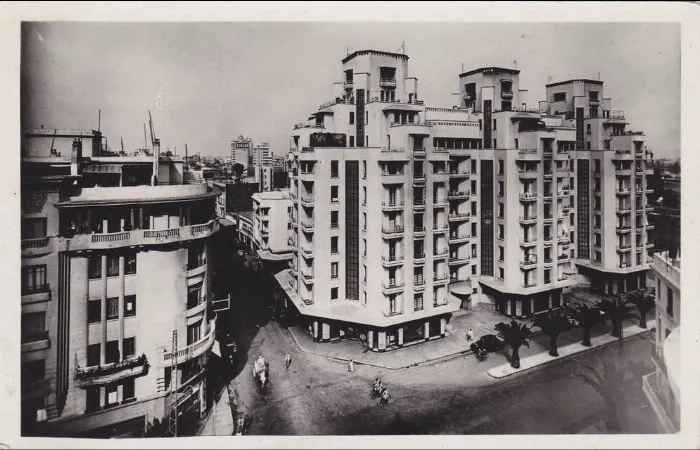 Старая фотография: дом Ассаяг с тремя башнями уступами на углу, пальмы, автомобиль и велосипедисты